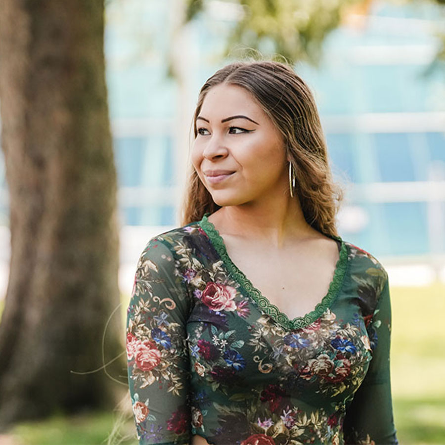 A college student with long hair poses for a portrait looking off to the side outside on a college campus.