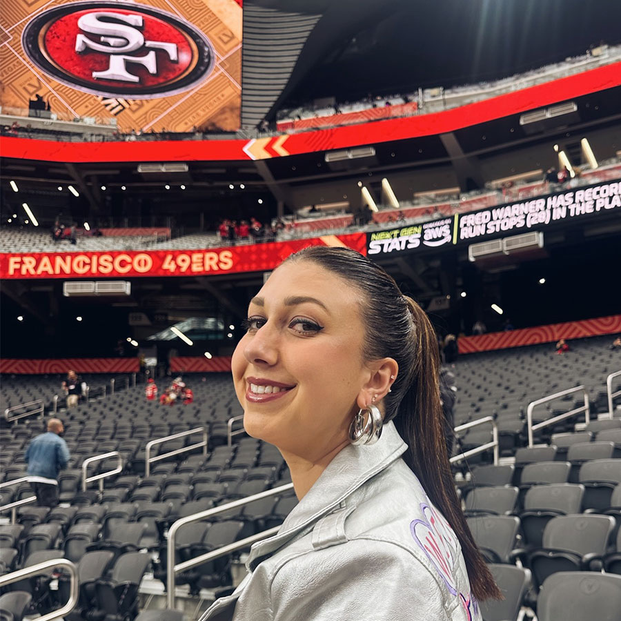 A woman in a silver jacket smiles at the camera inside of a large stadium where a screen displays the logo of the San Francisco 49ers football team.