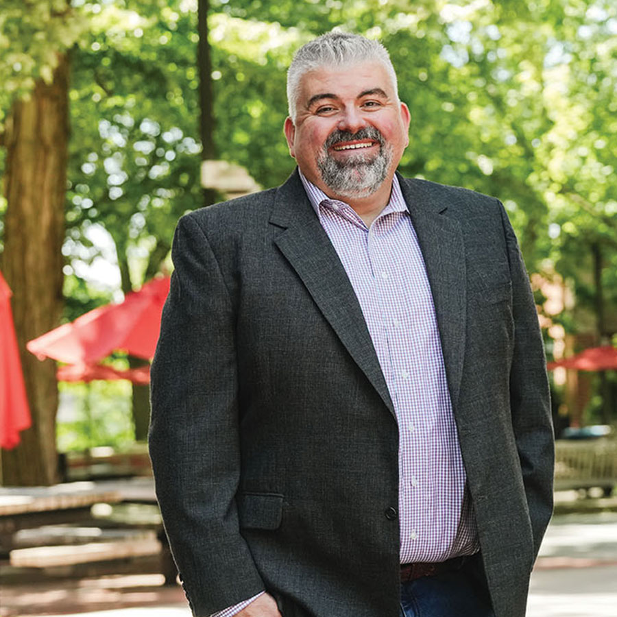 A man in a suit smiles at the camera outdoors in a college plaza.