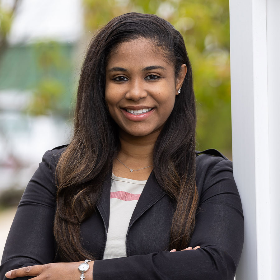 A woman in a black pantsuit stands outside, leaning on a white fence, and smiles at the camera