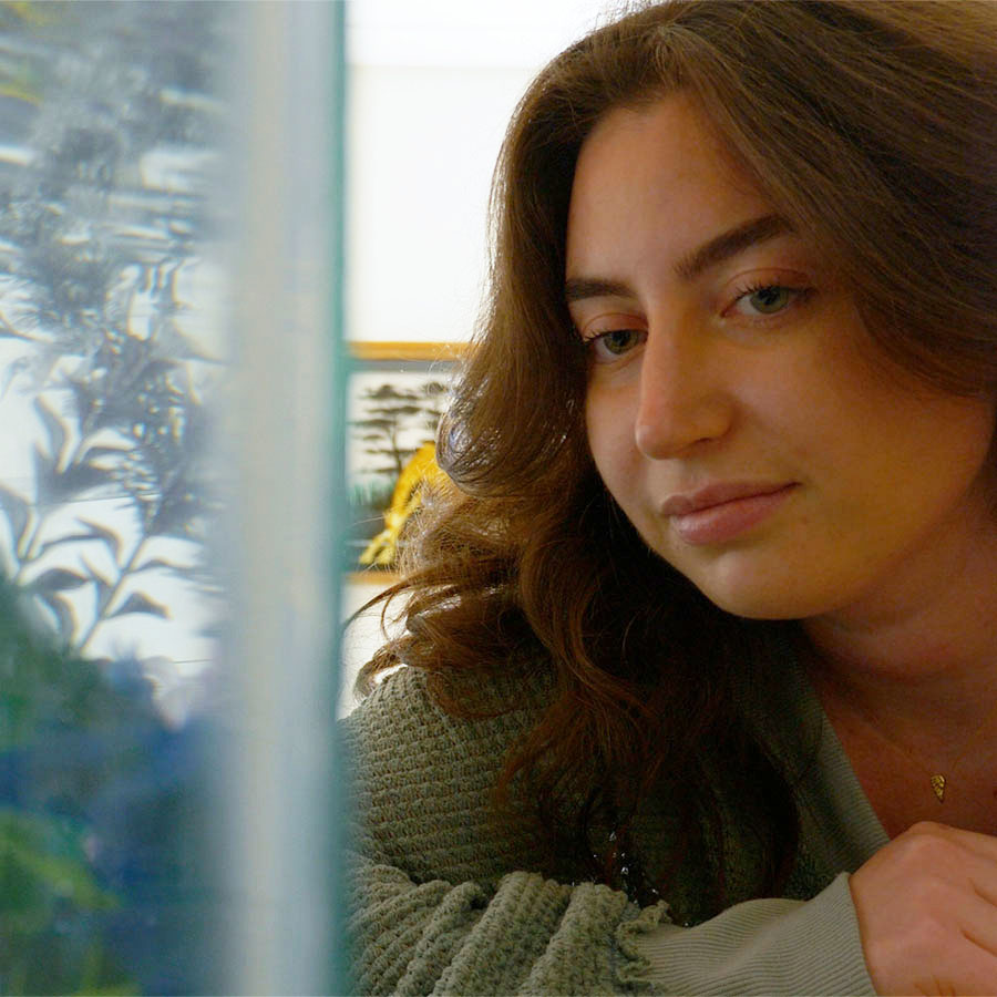 A college student with long brown hair looks thoughtfully at an aquarium tank in a lab.