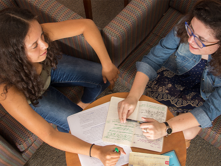 Viewed from above, two students, seated in armchairs, talk over a table covered with notebooks and paper. The students each hold a pen in one hand.