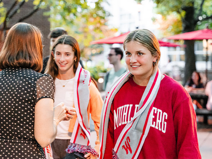 College students wearing Muhlenberg College gear receive and wear gray and red scarfs around their necks.