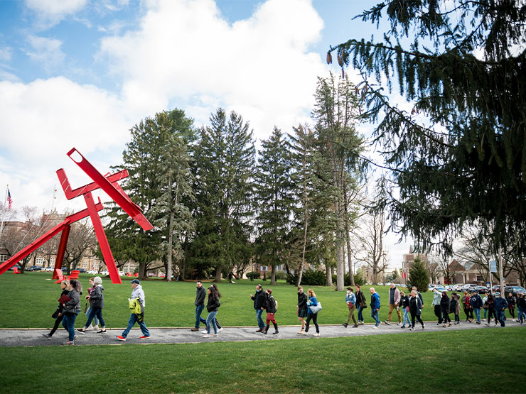 Clusters of families with young adults walk along a walkway on Muhlenberg College campus near the red modern sculpture Victors Lament.