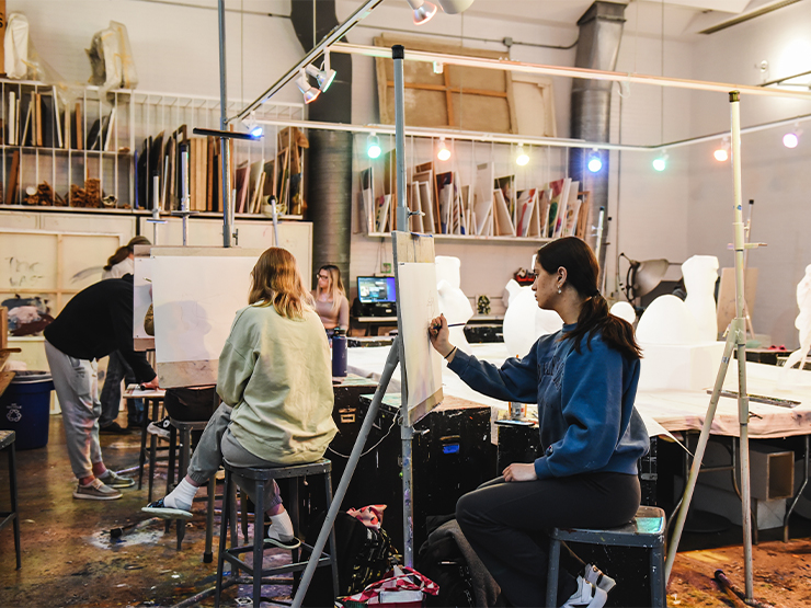 A group of students sit on stools as they put paint to canvases in an art studio.