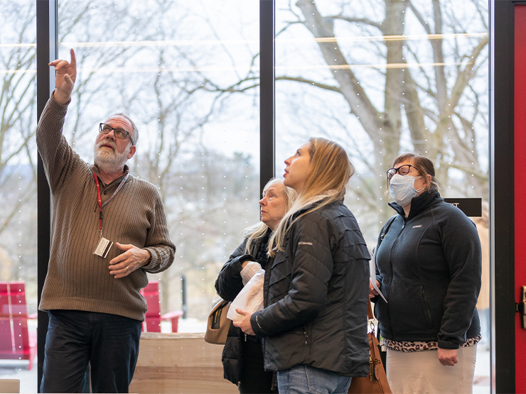 A man, speaking with a small group of people inside of a building, points up as he talks.