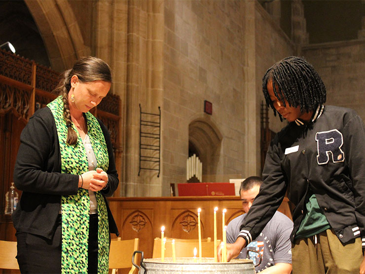 Inside, Egner Chapel, a college chaplain bows her heard while a student places a candle in a basin with others.