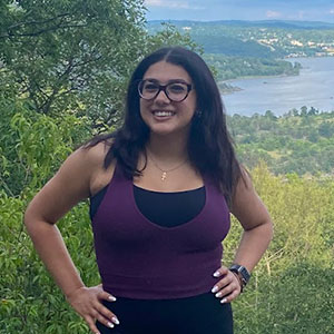 A college student in a tank top smiles, hands on hips, while posing atop a vista with a sprawling view.