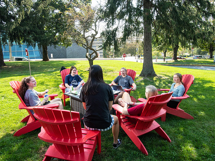 A group of young adults sit outside in a circle of red Adirondack chairs on a college green.