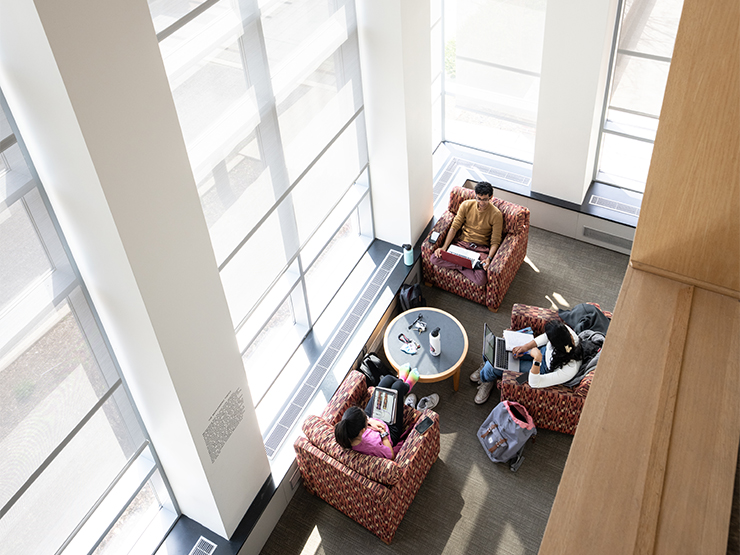 Seen from above, a cluster of students sit in large chairs around a small table, laptops on their laps and bags at their feet.