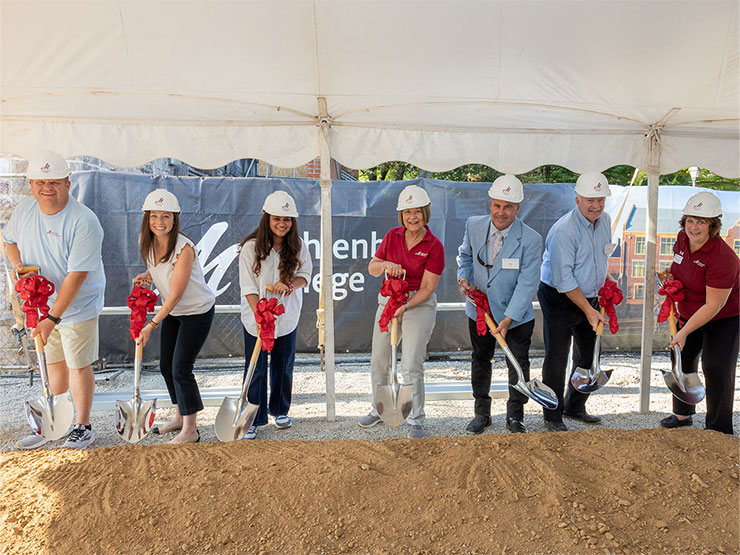 A group of college students, staff and officials smile while standing with shiny shovels festooned with ribbons, poised to dig into loose dirt beneath a white tent.