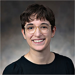 A person with short hair and glasses, wearing a black shirt, smiles at the camera in front of a grey backdrop.