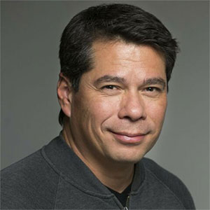 A man in a grey pullover smiles at the camera in front of a dark grey backdrop.