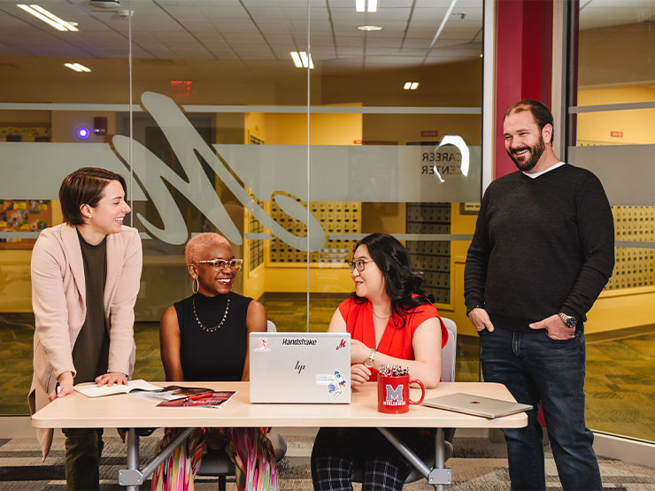 A college student and staff member sit at a desk with an open laptop while two other instructors stand on either side of the desk, all smiling.