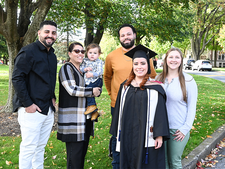 A recent graduate in formal commencement regalia stands outside on a college campus with family members and friends.