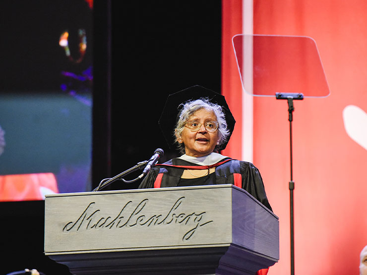 A person in commencement regalia speaks at a podium that is carved with the word 