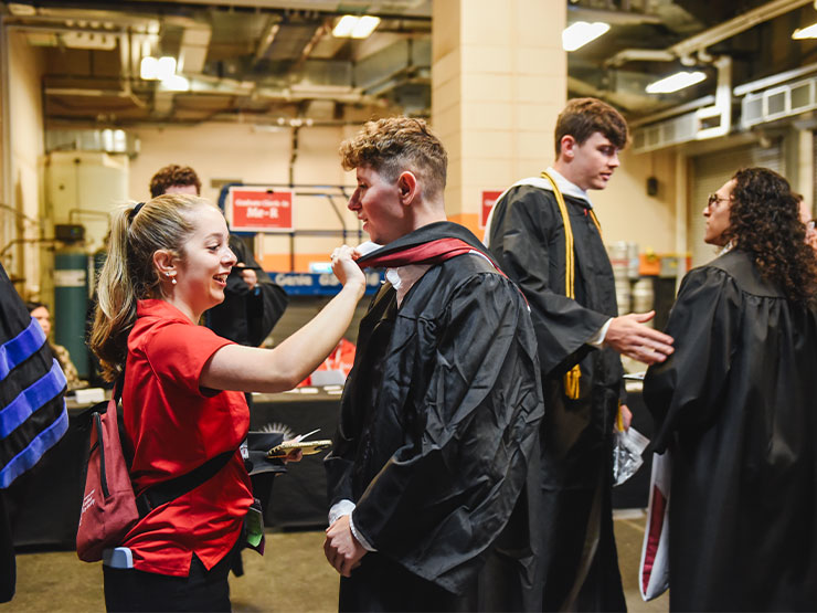 College students help one another adjust graduation regalia backstage at a commencement ceremony.