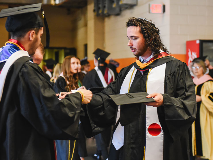 A pair of students dressed in graduation regalia connect backstage at an arena before the start of a ceremony.