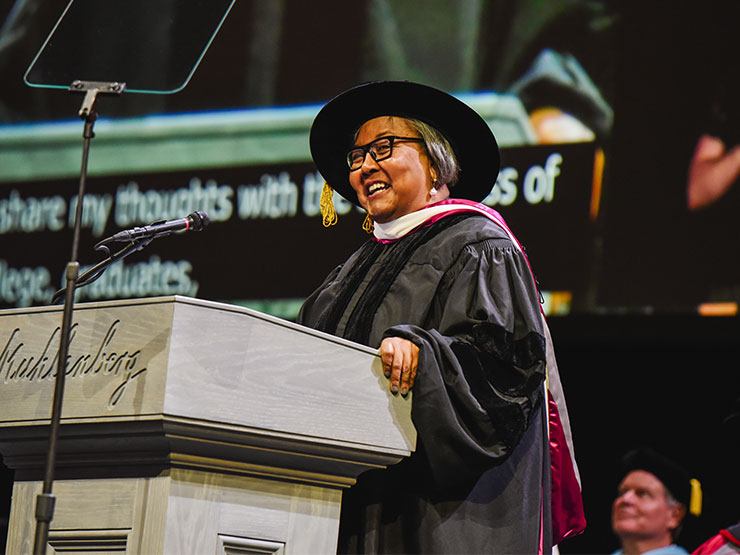 A woman in graduation regalia smiles while speaking at a podium.