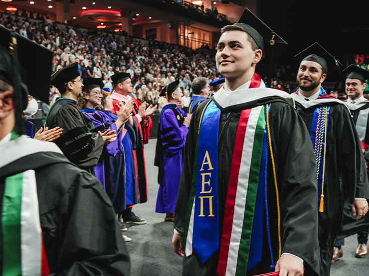 Students dressed in graduation regalia smile and walk in a line as onlookers applaud.