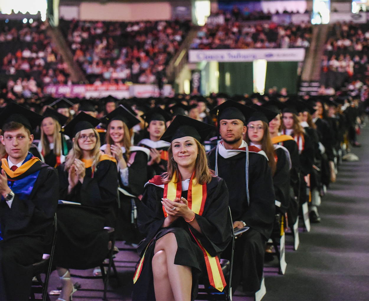 Students clad in full commencement regalia are clapping while seated in an auditorium