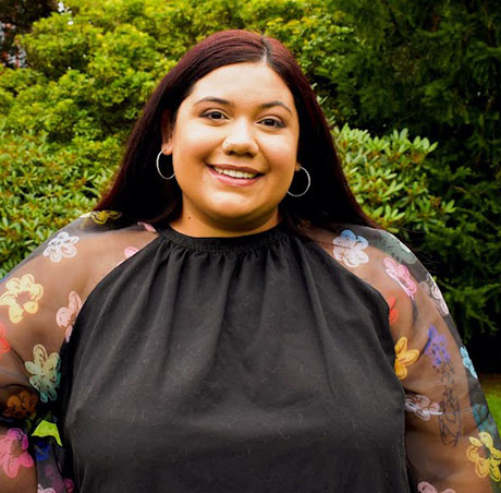 Office of Multicultural Life staff member Alyssa Rodriguez standing outdoors in front of green foliage.