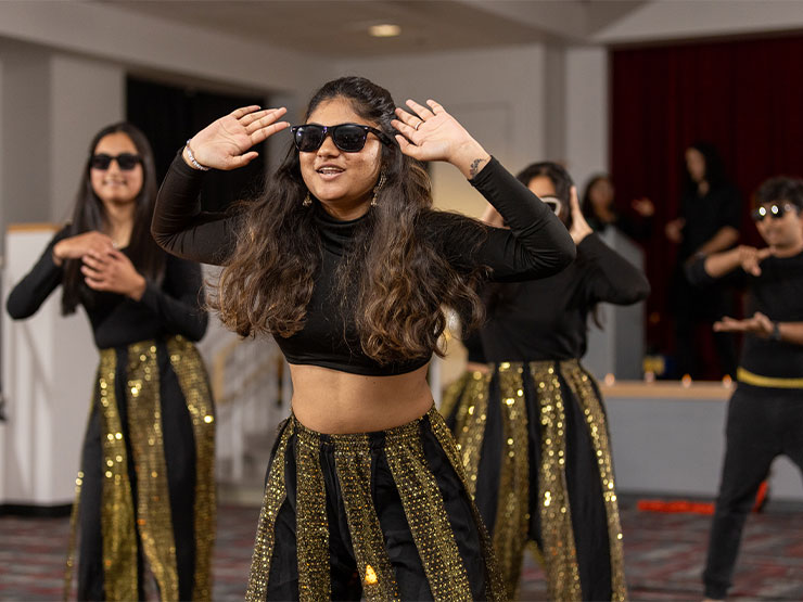 A group of college students wearing sunglasses, black tops and sparking gold and black pants perform a choreographed dance.