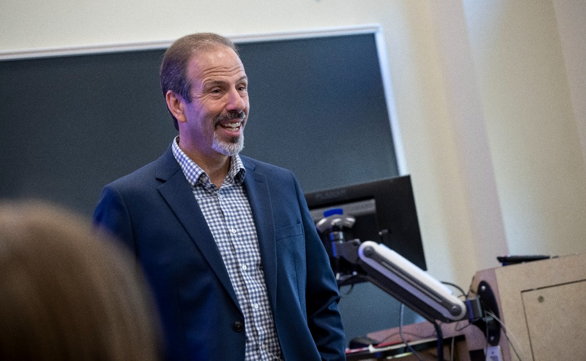 A smiling, male professor stands in front of a blackboard wearing a blue sports jacket.