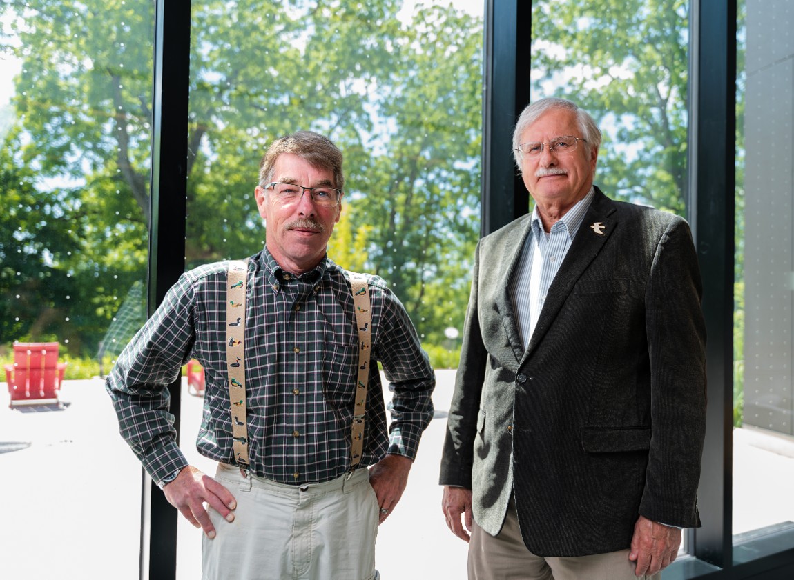 Two men stand facing the camera in front of a large window featuring strips of dots to prevent birds from crashing into it.