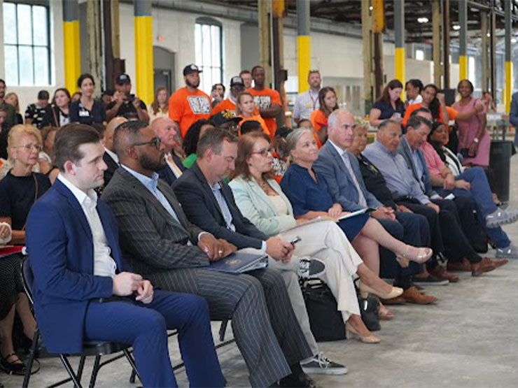 A large group of people, dressed in business clothes, sit in folding chairs inside of an industrial building.