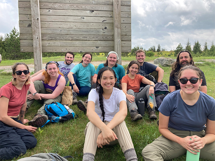 A group of students dressed in hiking gear smile at the camera while taking a break on a grassy hill in a mountainous region..