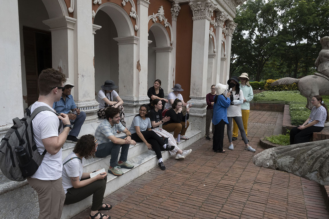 A group of people sit on the steps of a brick building with stone columns in Sonargaon, the old capital of Bangladesh