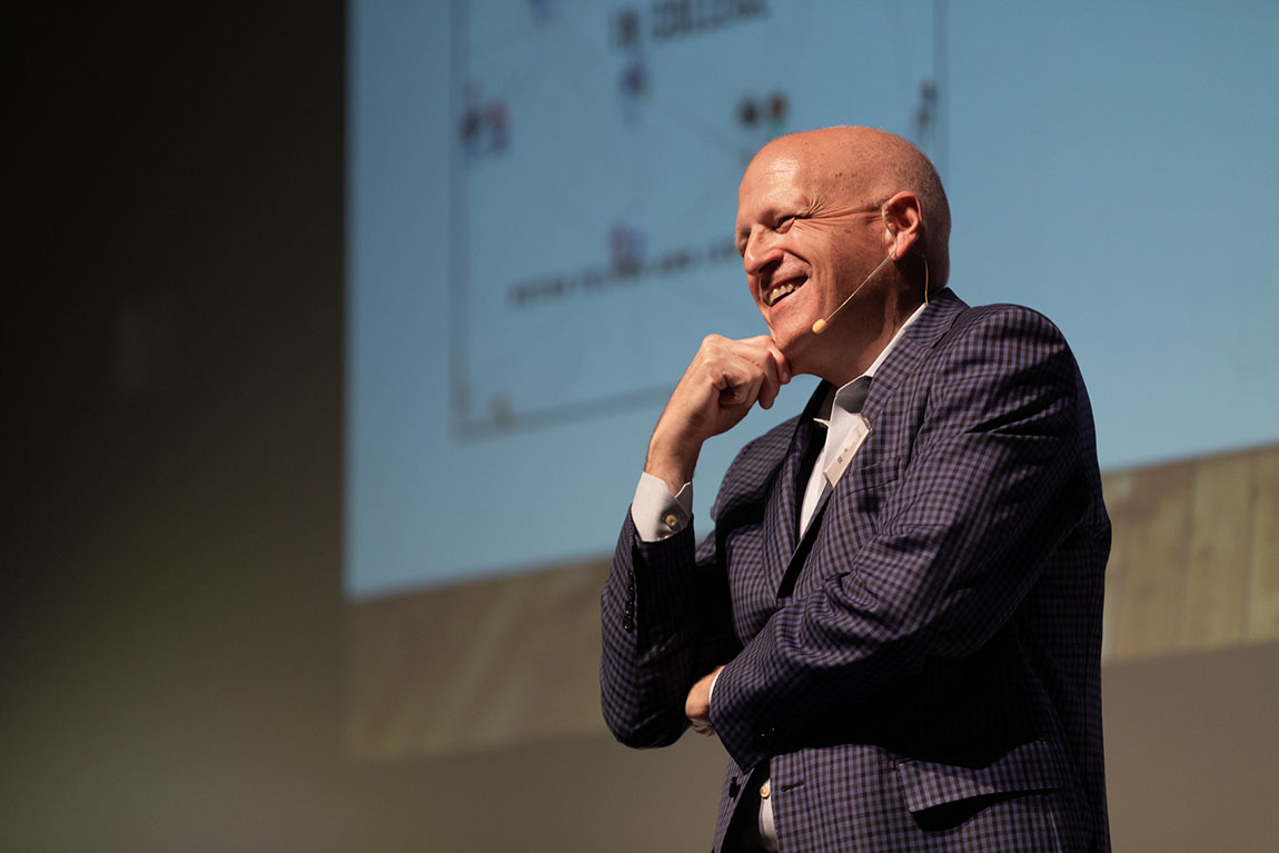 A bald man wearing a microphone smiles in front of a screen with some words projected on it