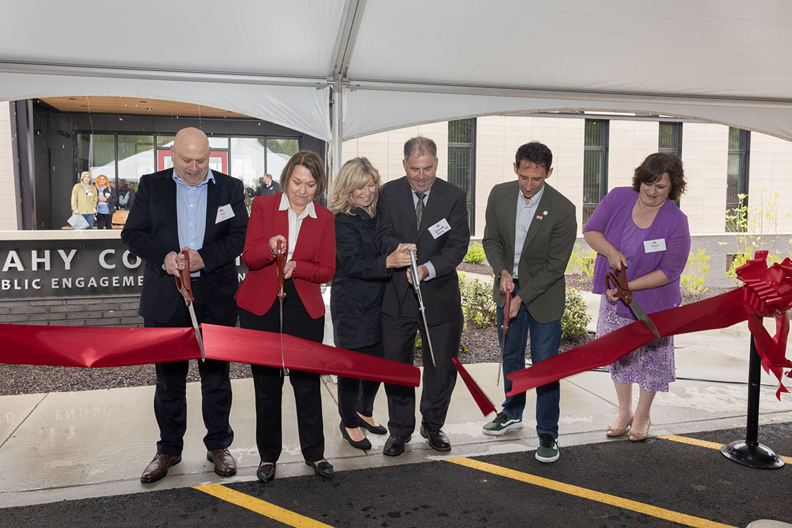 A group of six people uses giant scissors to cut a red ribbon in front of a new building