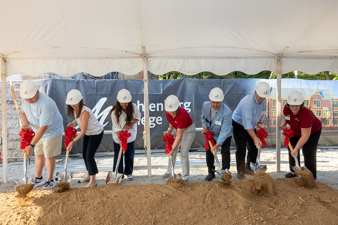 A group of seven people wearing white hard hats dig in dirt using shovels with red bows on them