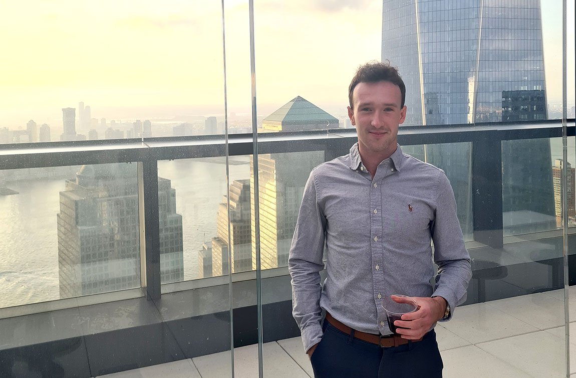 A recent college graduate in a button-down shirt stands on a balcony with a city skyline in the background