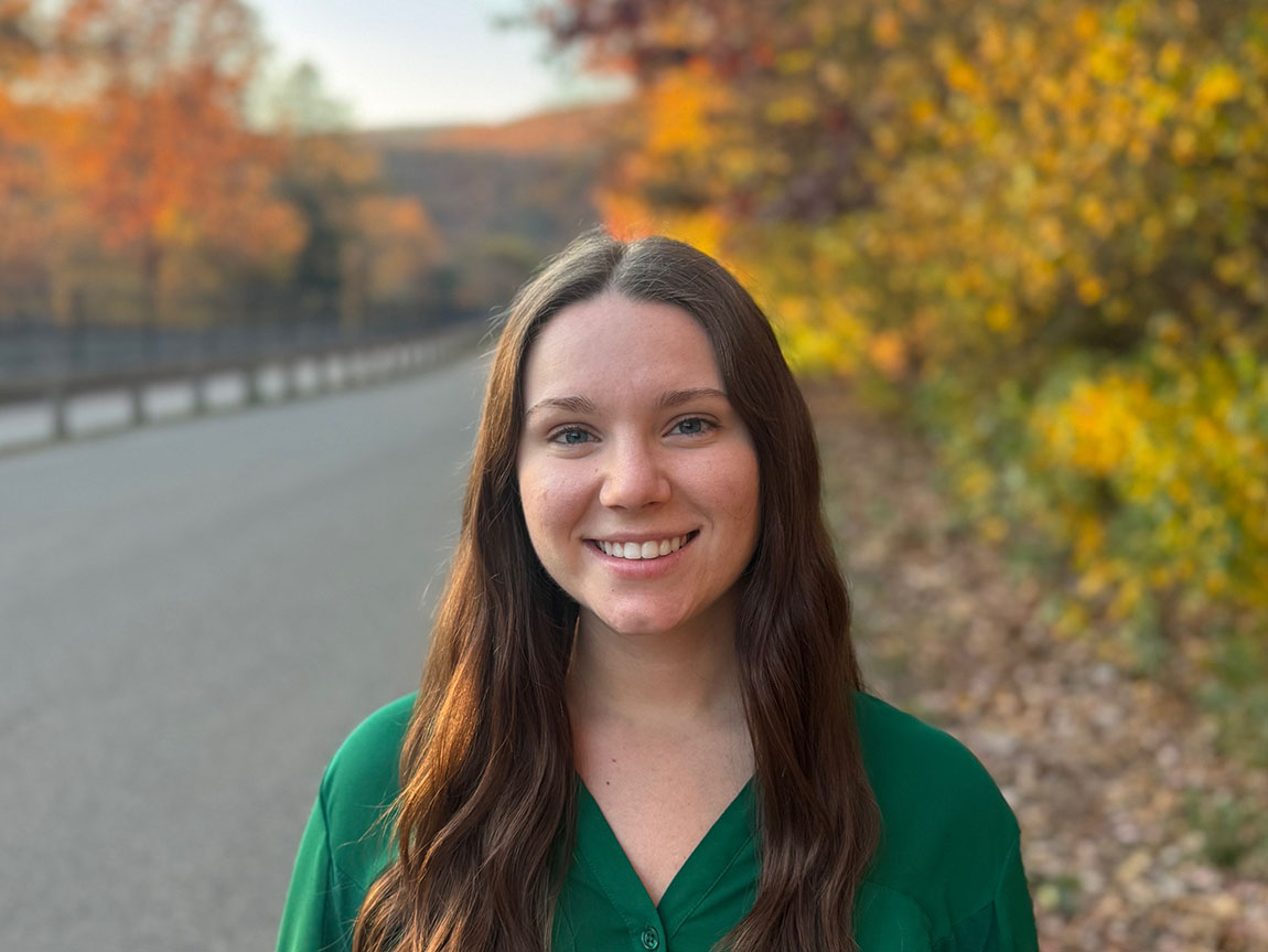 A young person in a green shirt smiles for a photo outside with fall foliage in the background