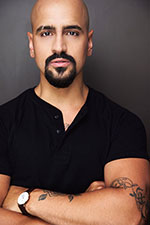 A headshot of an actor who is bald and has dark facial hair in a black T-shirt on a gray background