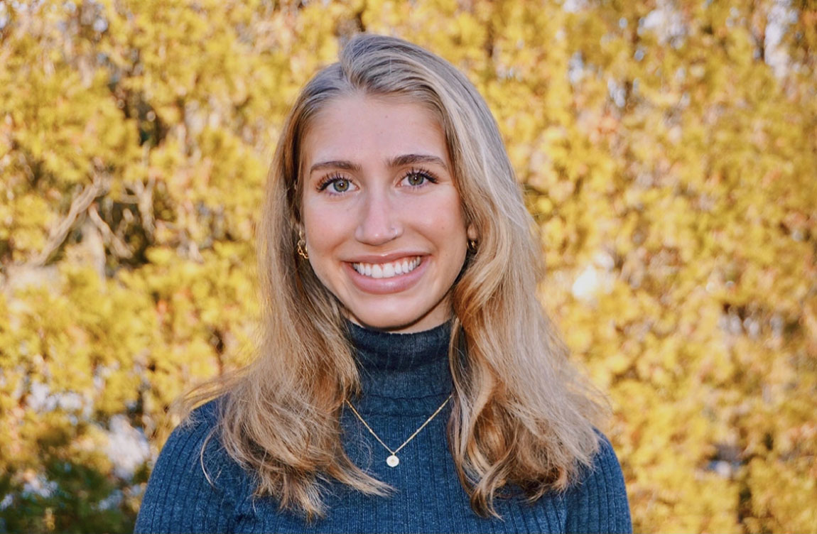 A headshot of a young professional with long blonde hair smiling at the camera in front of a yellow background