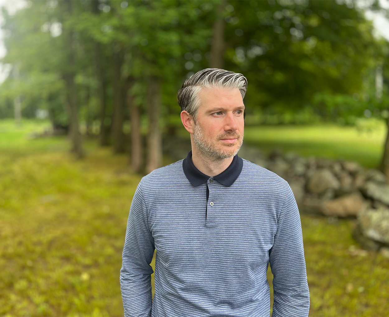 A man stands outdoors in a setting surrounded by trees while looking off camera.