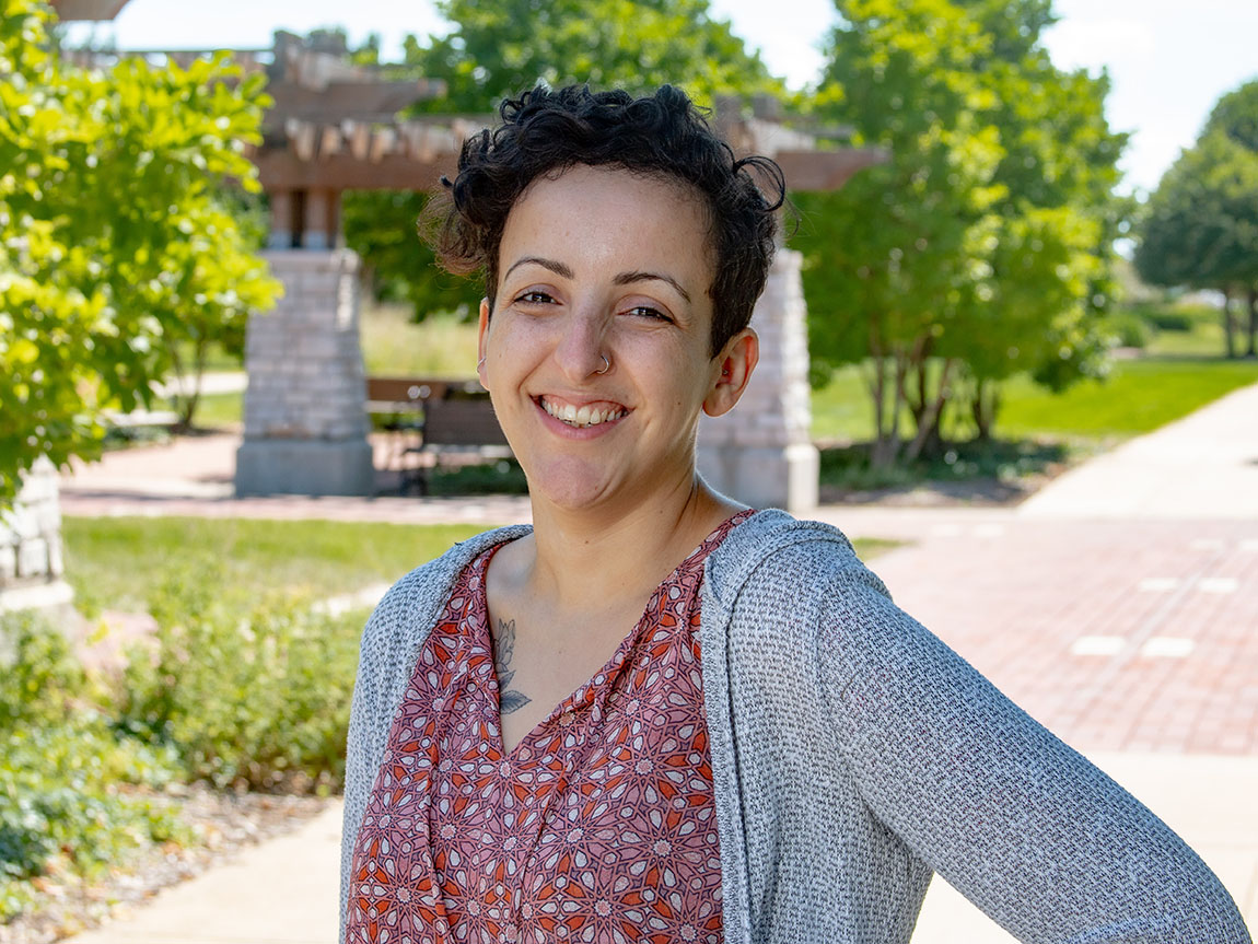 A headshot taken outside of Hannah Mesouani, who has short dark hair and is wearing a flowered top and gray sweater