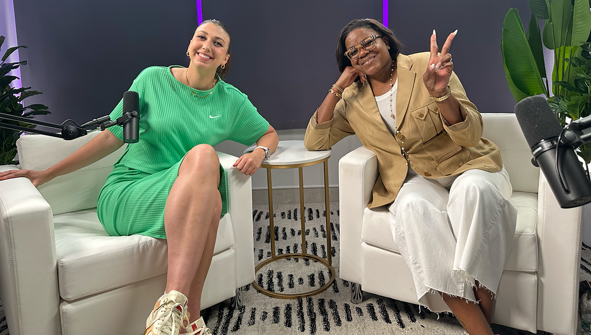 A young adult in a green dress smiles next to an older adult in a tan blazer flashing a peace sign