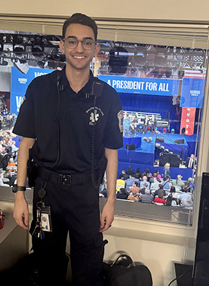 A college student in an EMS uniform stands in front of a window overlooking a rally stage