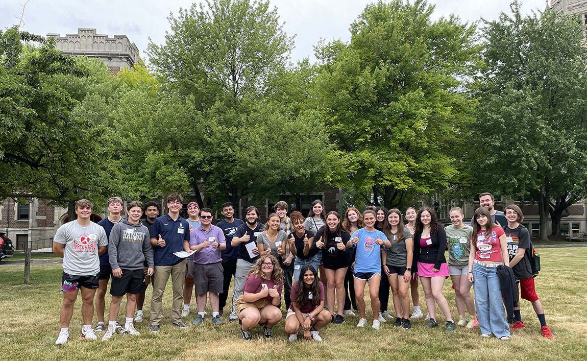 A large group of incoming college students stand in a row and smile while two college students and matching maroon T-shirts sit and smile in front of them on a lawn with trees in the back