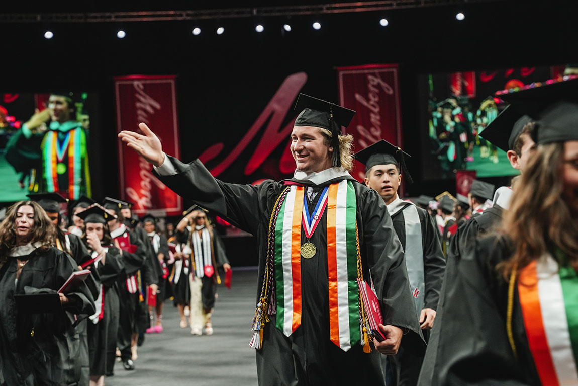 A college graduate in regalia gestures to the crowd and smiles while leaving commencement