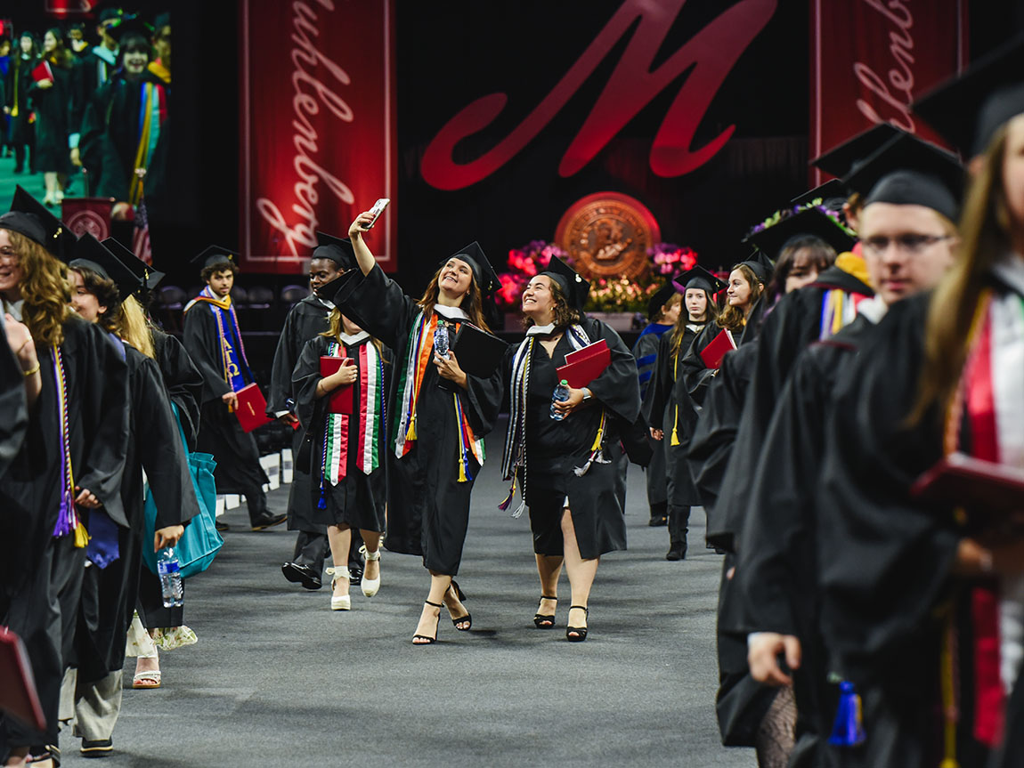 Two college graduates process out of Commencement while taking a selfie on their phone