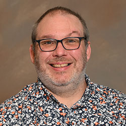 A headshot of a college professor with glasses, a gray beard, and a patterned button up shirt on