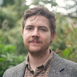 A college professor with brown hair and beard and a blazer on photographed outside