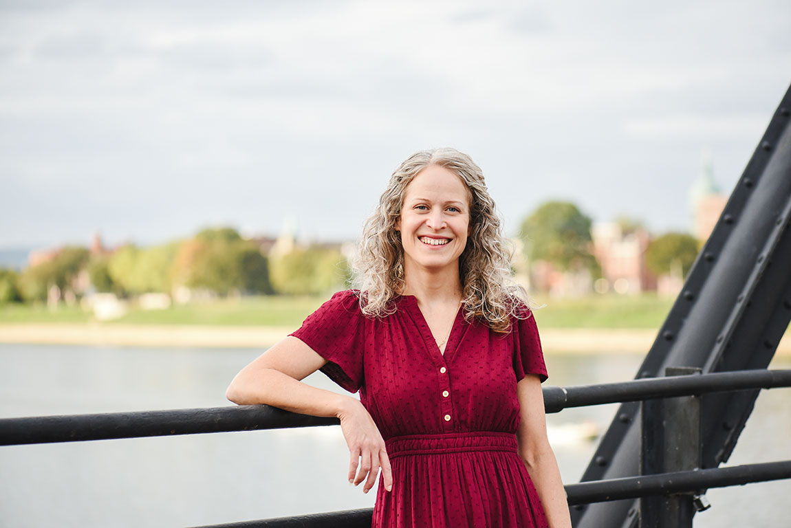 A woman with shoulder-length curly hair wearing a red dress smiles at the camera as she stands atop a bridge overlooking a river