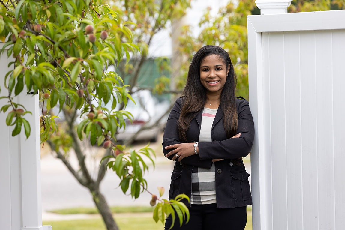 A woman in a black pantsuit stands outside, leaning on a white fence, and smiles at the camera